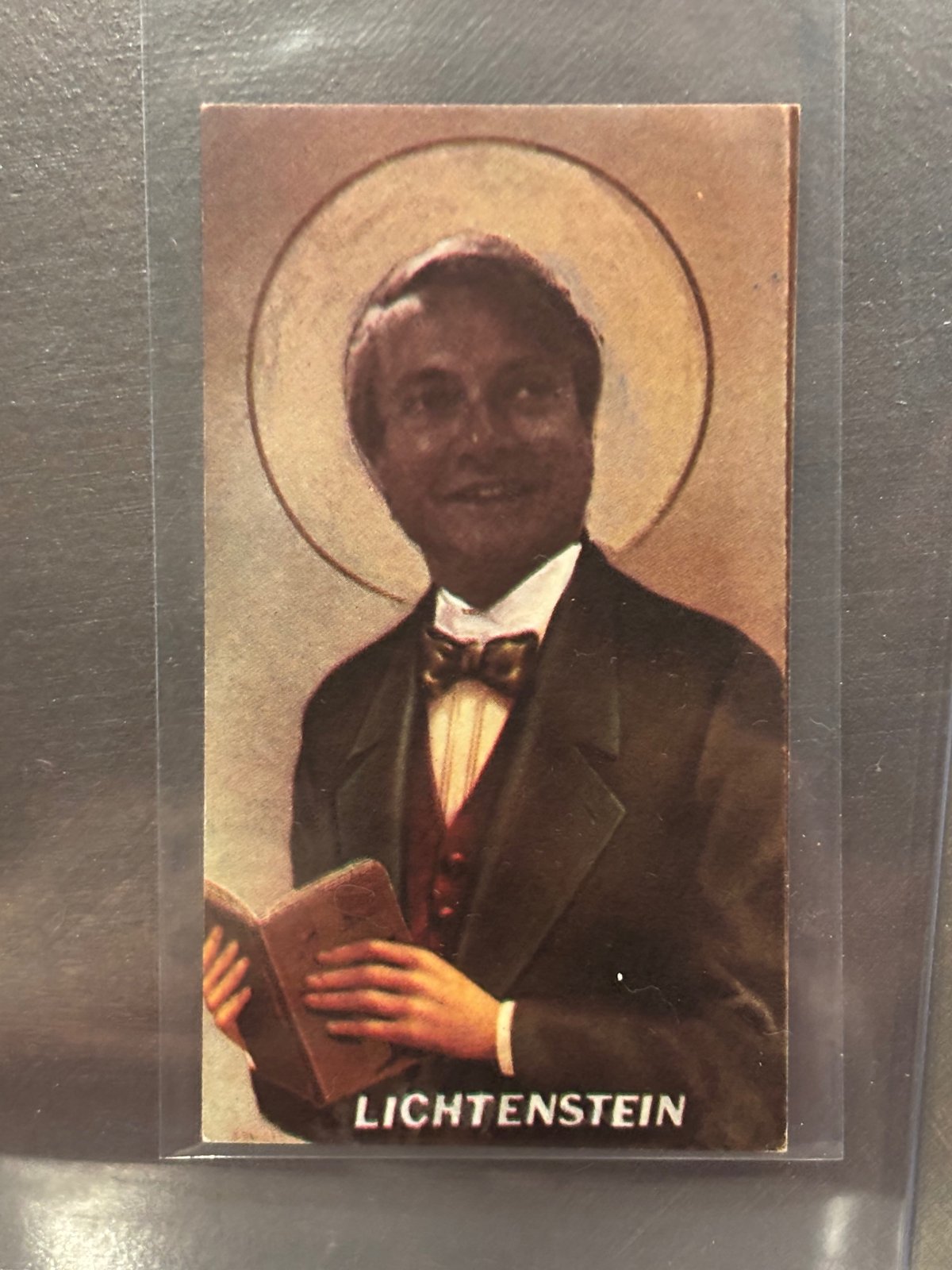 Holy Holy Art Card of Roy Lichtenstein as a haloed saint in a bow tie holding a book, captioned "LICHTENSTEIN."